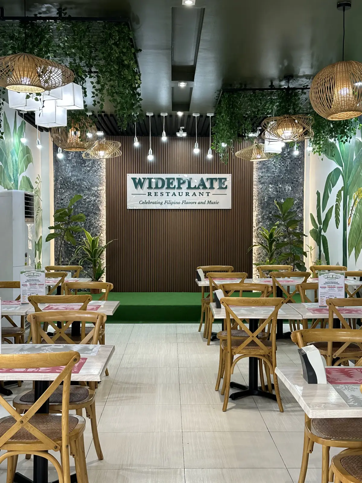Wideplate dining room: banana-leaf murals, rattan pendant lights, and the wordmark on a wood-slat wall